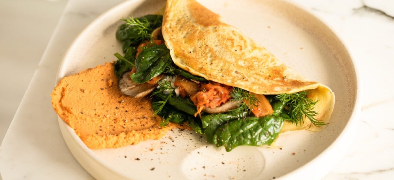 A folded omelette filled with fresh greens, mushrooms, and herbs, served with a dollop of reddish-orange spread on a light-colored plate.